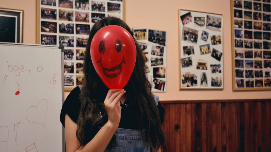 A young woman holding a red balloon with a cartoon face, standing in a photo-filled room.